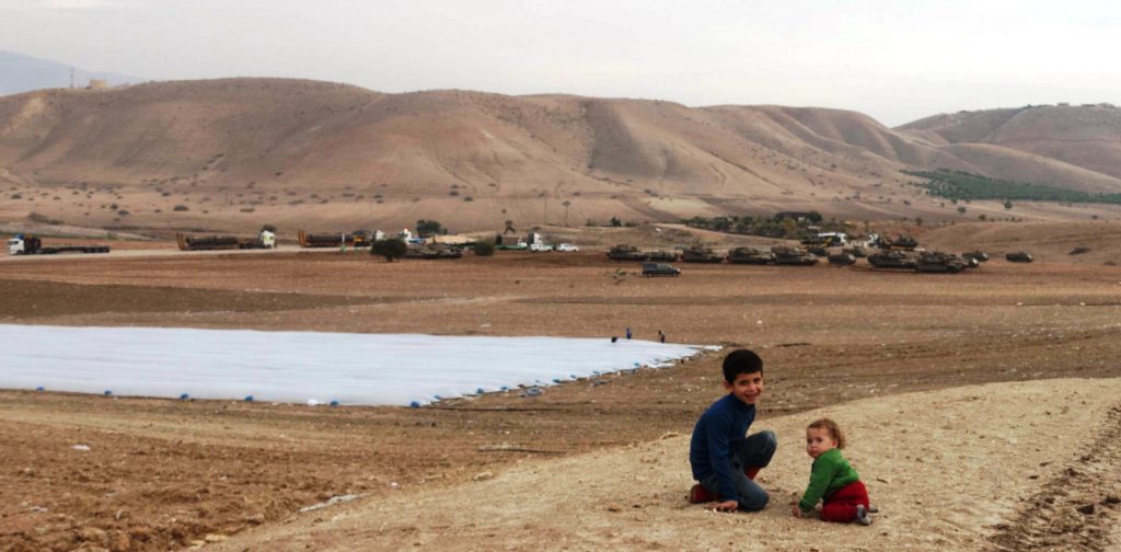 Ein Bild aus dem wenige Kilometer entfernten Farisiya veranschaulicht die Problematik: während die Erwachsenen auf den Feldern arbeiten und die Kinder spielen ziehen im Hintergrund Panzer zu militärischen Übungen auf; ©EAPPI